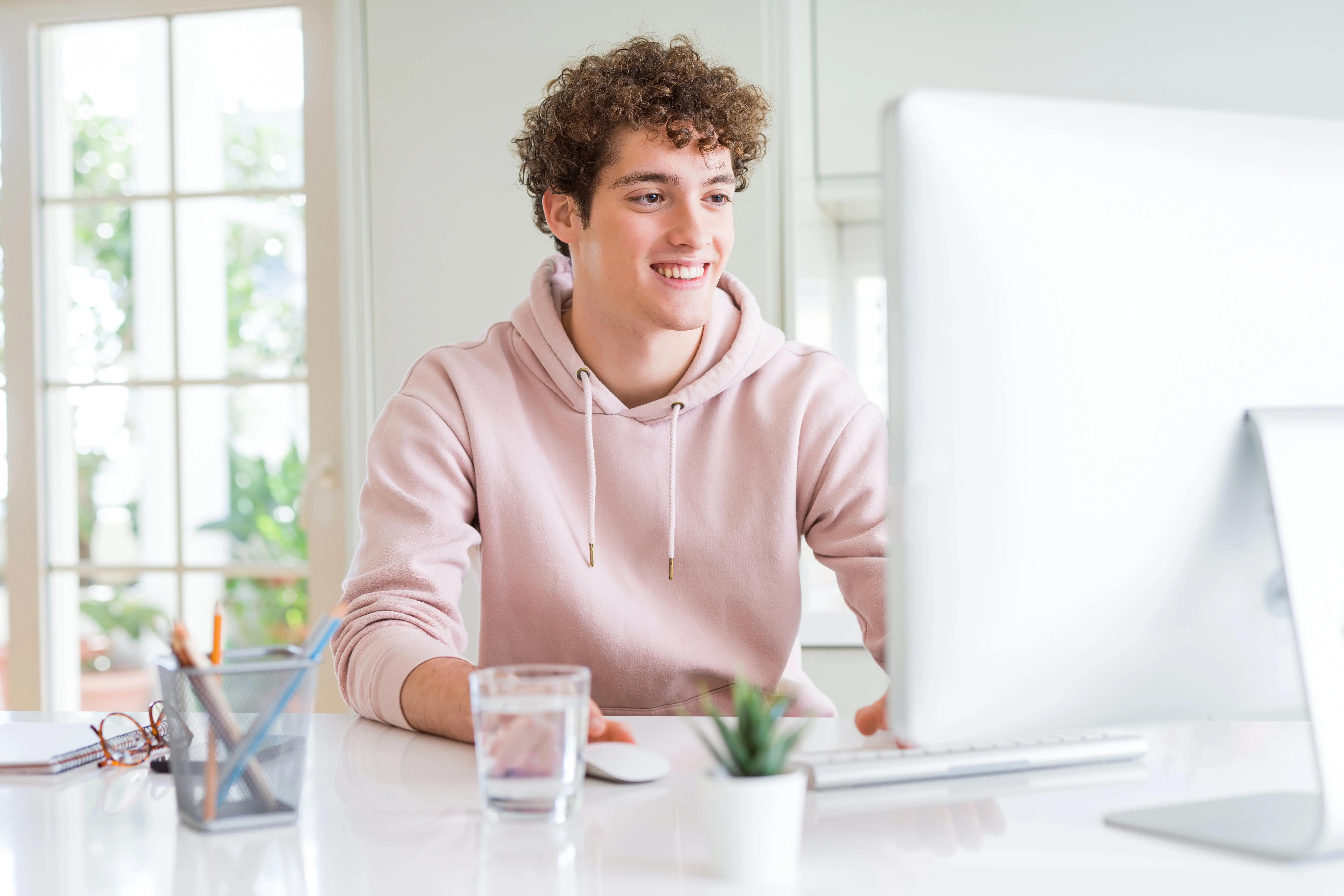 Młody chłopak siedzi przy komputerze, rozmawia online. Jest ubrany w różową bluzę.